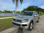 Silver Volkswagen SUV parked near a lake with palm trees in the background, showcasing its low miles and great condition.