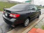 2012 Toyota Corolla LE showing rear view, black color, parked on the street with wet pavement.