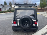 Rear view of a 1999 Jeep Wrangler Sahara with off-road tires and a luggage rack parked on a street.