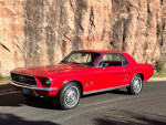 1967 Ford Mustang in candy apple red parked on a scenic road with rocky background.