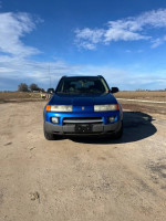 Front view of a blue 2004 Saturn VUE SUV parked on a dirt road under a cloudy sky.